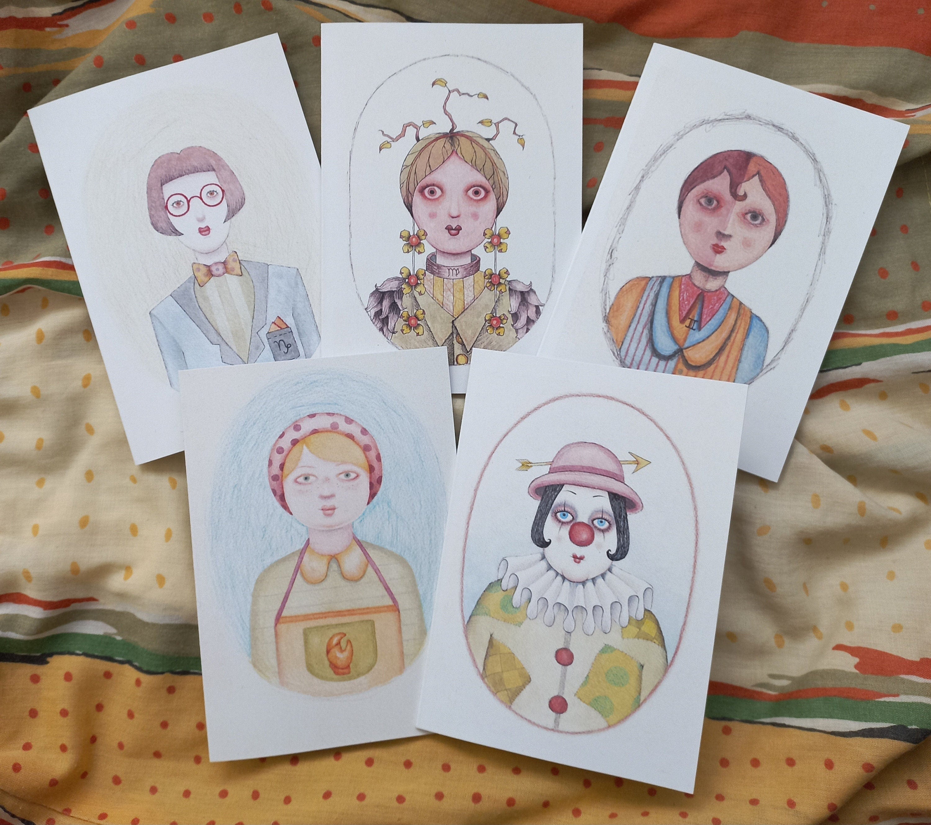 Five illustrated astrology portraits on white greeting cards laid on a colourful fabric background