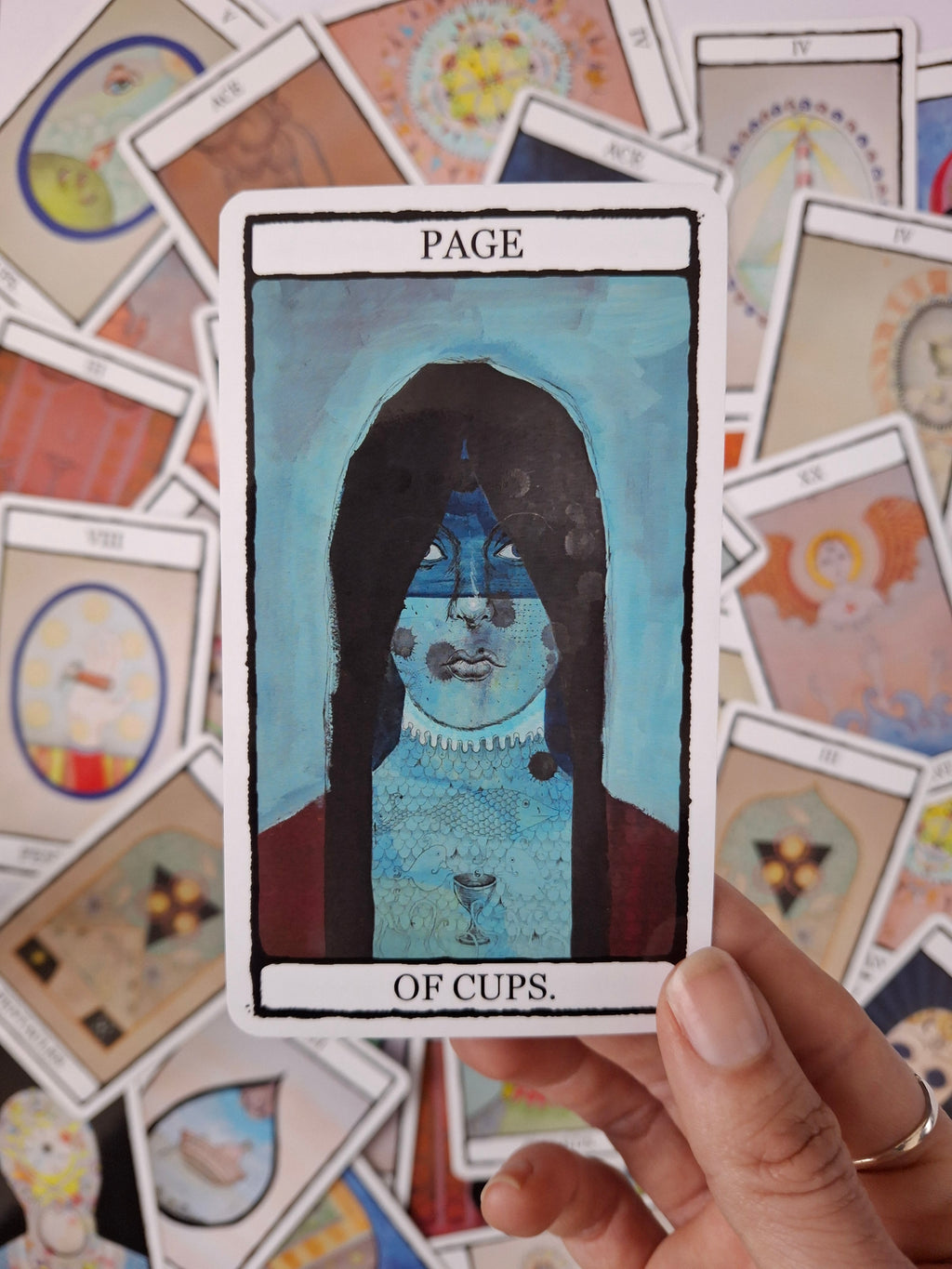 Page of Cups card from a tarot deck featuring an illustration of a dark haired girl with a blue background